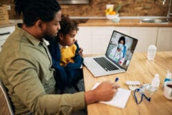 Black father and daughter communicating with a doctor via video call from home during COVID-19 pandemic. Focus is on girl.