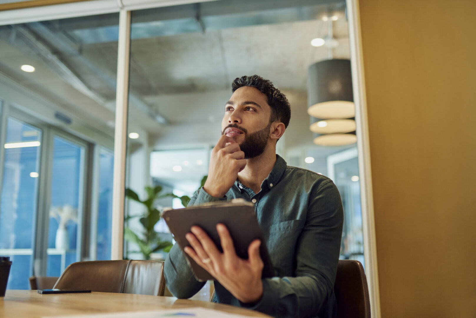 An image of a young businessman sitting in a modern office, thoughtfully analyzing data on his tablet while contemplating his next strategic move.