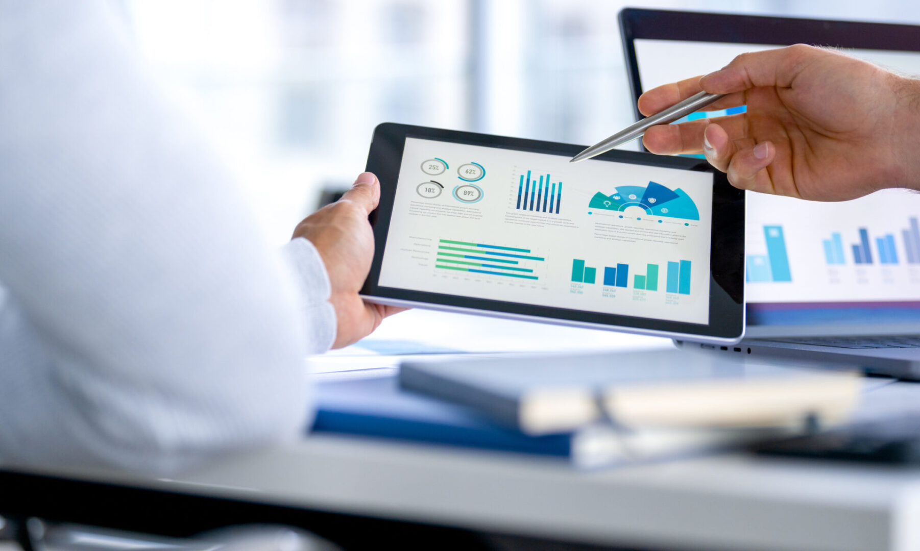 Two business men meeting and looking at financial data, charts and graphs on a digital tablet and a laptop computer. Two business men meeting and looking at financial data, charts and graphs on a digital tablet and a laptop computer. There are documents and paperwork on the table. One man is pointing at the screen
