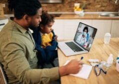 Black father and daughter communicating with a doctor via video call from home during COVID-19 pandemic. Focus is on girl.