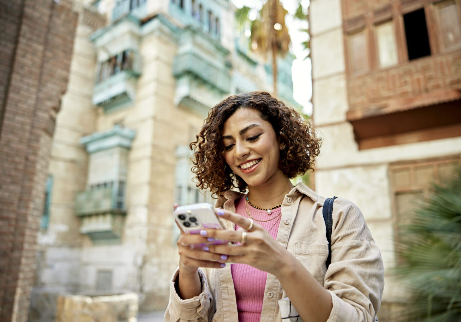 Waist-up view of casually dressed woman with curly brown hair looking at smart phone and smiling while visiting historic Al-Balad, Jeddah.