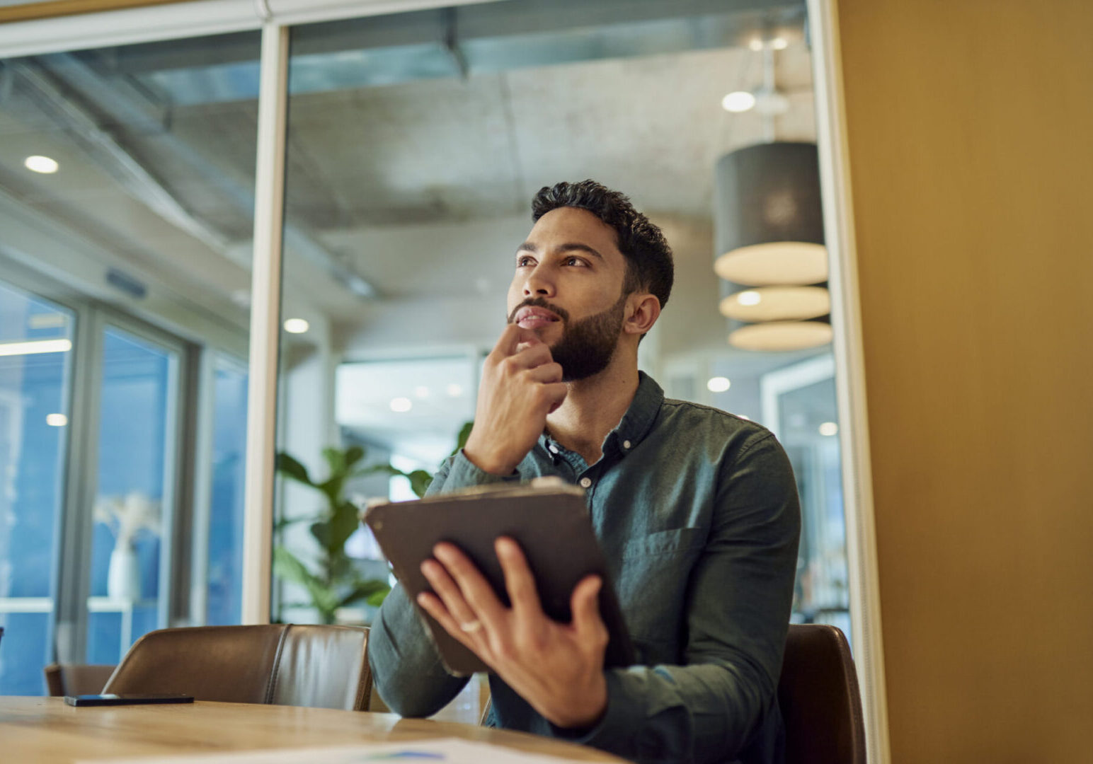 An image of a young businessman sitting in a modern office, thoughtfully analyzing data on his tablet while contemplating his next strategic move.